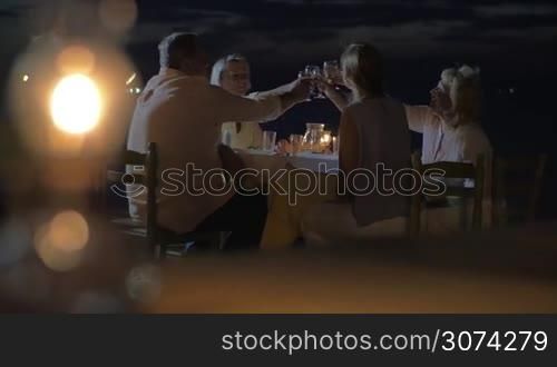Big united family gathered round the table outdoor at night. They are clinking wine glasses and talking.
