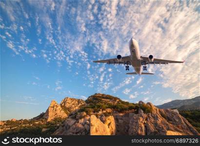 Big white airplane is flying over rocks at sunrise. Landscape with passenger airplane, mountains, colorful blue sky with clouds. Passenger aircraft is landing. Business travel. Commercial plane.