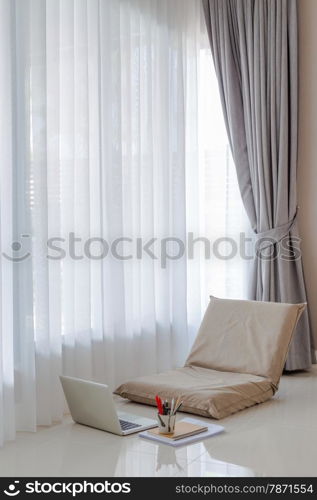 Big window of living room with sun light come through thin white curtain. Temporary area for working at home with foldable lazy chair, laptop, documents and stationery on the floor.