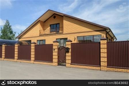 Big yellow brick cottage with balcony and brown metal fence, sunny day