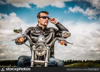 Biker man wearing a leather jacket and sunglasses sitting on his motorcycle looking at the sunset.