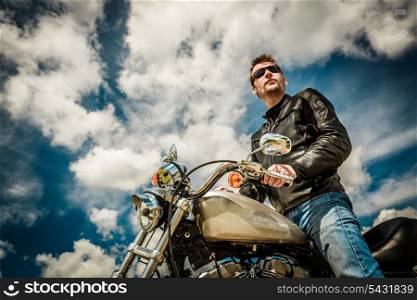 Biker man wearing a leather jacket and sunglasses sitting on his motorcycle looking at the sunset.
