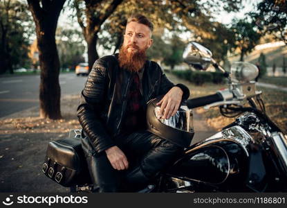 Biker with helmet in hands sitting on the ground against his chopper. Vintage bike, rider and his motorcycle, freedom lifestyle, biking. Biker sitting on the ground against his chopper