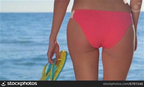 Bikini girl with flip flops in hand walking to the sea rear view