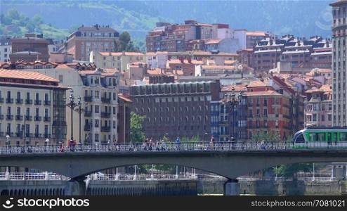 Bilbao Tram travels across bridge with city behind it