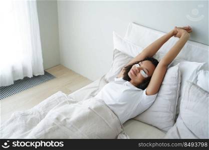 Biracial woman stretching in bed after wake up with moisturizing under eye patches on face. Young girl lying in bedroom with facial cosmetic product on undereye skin in the morning. Skincare treatment. Young girl stretching in bed after wake up with under eye patches on face. Skincare treatment