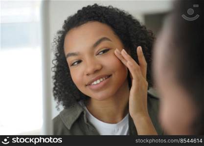 Biracial young girl looks at mirror, touches delicate under eye skin, feels happy after moisturizing mask. Mixed race woman apply daily facial cream. Natural beauty and skin care concept.. Biracial young girl looks at mirror, applying moisturizing facial cream. Natural beauty, skincare
