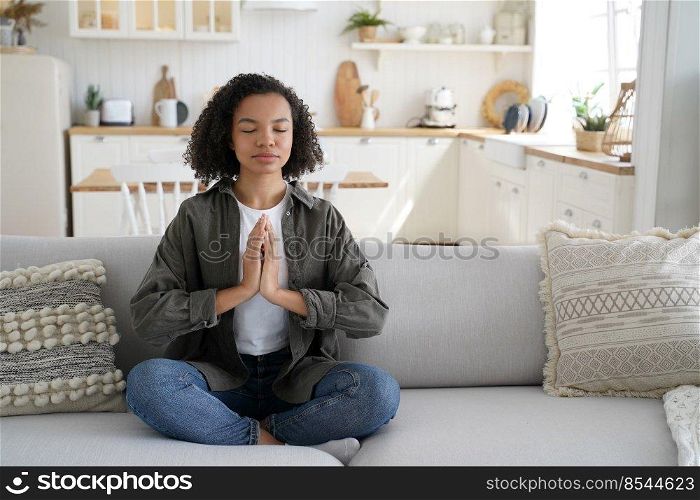 Biracial young girl practicing yoga doing Padmasana exercise at home, calm relaxed mixed race woman sitting in lotus pose on sofa, meditating in living room. Healthy lifestyle habbit, wellness.. Biracial woman practices yoga at home, sitting in lotus pose on sofa. Meditation, healthy lifestyle