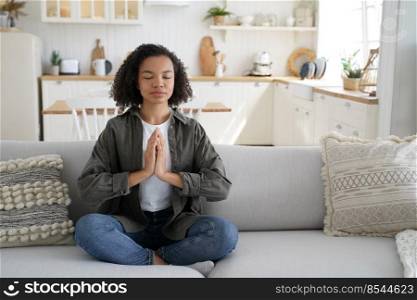 Biracial young girl practicing yoga doing Padmasana exercise at home, calm relaxed mixed race woman sitting in lotus pose on sofa, meditating in living room. Healthy lifestyle habbit, wellness.. Biracial woman practices yoga at home, sitting in lotus pose on sofa. Meditation, healthy lifestyle