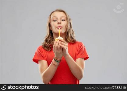 birthday and people concept - teenage girl in red t-shirt holding cupcake with one burning candle and blowing it out over grey background. teenage girl with one candle on birthday cupcake
