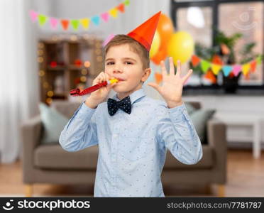 birthday, celebration and childhood concept - portrait of smiling little boy in party hat with blower having fun over decorated home room background. smiling boy in birthday party hat with blower