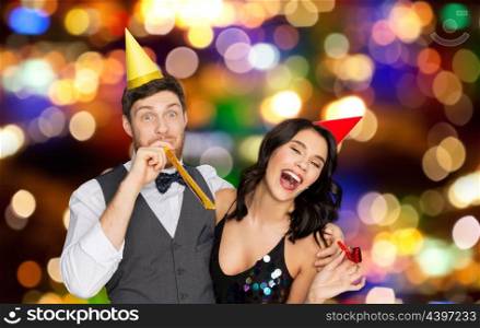 birthday, celebration and holidays concept - happy couple with party blowers and caps having fun over festive lights background. happy couple with party blowers having fun. happy couple with party blowers having fun