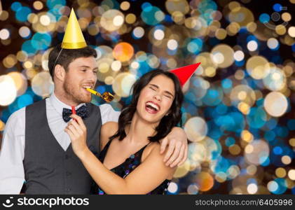 birthday, celebration and holidays concept - happy couple with party blowers and caps having fun over festive lights background. happy couple with party blowers having fun