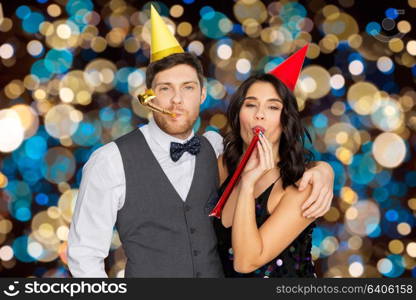 birthday, celebration and holidays concept - happy couple with party blowers and caps having fun over festive lights background. happy couple with party blowers having fun