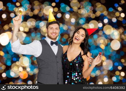 birthday, celebration and holidays concept - happy couple with party blowers and caps having fun over festive lights background. happy couple with party blowers having fun