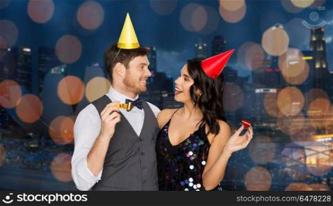 birthday, celebration and holidays concept - happy couple with party blowers and caps having fun over singapore city night lights background. happy couple with party blowers having fun. happy couple with party blowers having fun