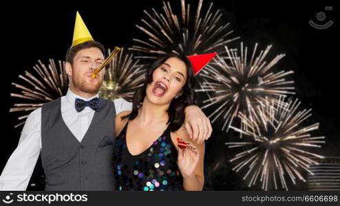 birthday, celebration and holidays concept - happy couple with party blowers and caps having fun over firework lights at night city background. happy couple with party blowers having fun