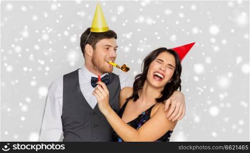 birthday, celebration and holidays concept - happy couple with party blowers and caps having fun over grey background and snow. happy couple with party blowers having fun