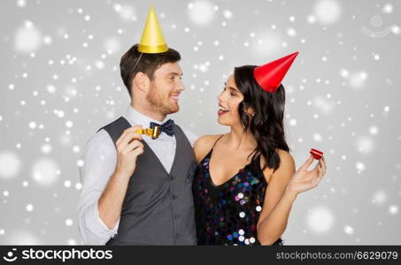 birthday, celebration and holidays concept - happy couple with party blowers and caps having fun over grey background and snow. happy couple with party blowers having fun