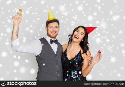 birthday, celebration and holidays concept - happy couple with party blowers and caps having fun over grey background and snow. happy couple with party blowers having fun