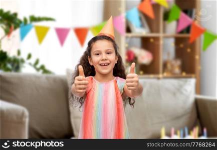 birthday, childhood and people concept - portrait of smiling little girl in dress and party hat showing thumbs up over cake and garland flags in decorated home room background. happy girl in birthday party hat showing thumbs up