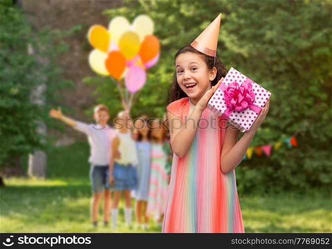 birthday, childhood and people concept - portrait of smiling little girl in dress and party hat with gift box over group of children at summer park background. smiling girl with gift on birthday party at park