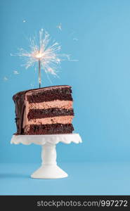 Birthday concept with a slice of chocolate layered cake with a sparkler on top, on a blue table. New year frame with piece of cake with a firework.