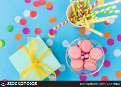 birthday party, celebration and decoration concept - gift box, pink macarons, paper straws and confetti on blue background. birthday gift, macarons and paper straws for party