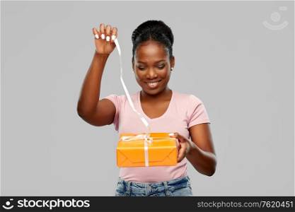 birthday present and surprise concept - happy african american young woman untying ribbon on gift box over grey background. happy african american woman with gift box