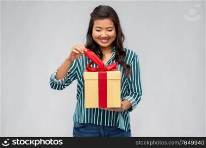 birthday present and surprise concept - happy asian young woman opening gift box with red bow over grey background. happy asian woman opening gift box