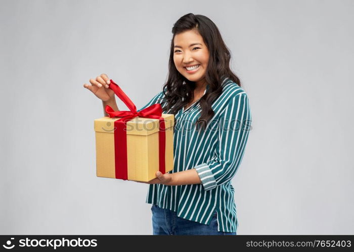 birthday present and surprise concept - happy asian young woman opening gift box with red bow over grey background. happy asian woman opening gift box