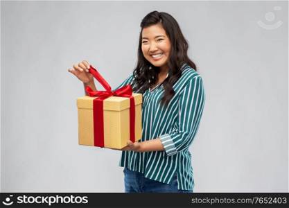 birthday present and surprise concept - happy asian young woman opening gift box with red bow over grey background. happy asian woman opening gift box