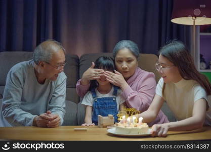 Birthday surprise concept. Happy Asian family grandparents and mother celebrating enjoy singing birthday song party with cake of granddaughter blowing candle at home in night time