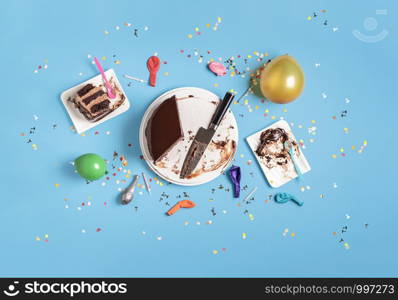 Birthday table frame with eaten chocolate layered cake, colorful balloons, and confetti on a blue background. Above view of new year celebration.