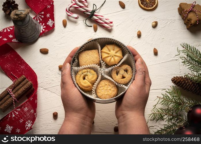 Biscuits box in woman's hands above view. White table decorated with Christmas sweets and ornaments in the background