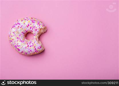 Bitten doughnut with colorful sprinkles and white glaze, on a pink paper background. Above view. Minimalist style image. Baked sweet pastry item.