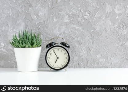 Black alarm clock and green succulent in white pot on table on background of gray concrete wall. Its five to eight a.m Concept good morning Copy space.. Black alarm clock and green succulent in white pot on table on background of gray concrete wall. Its five to eight a.m Concept good morning Copy space