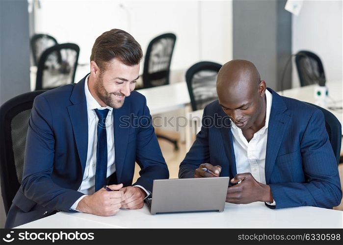 Black and caucasian smiling businessmen looking at a laptop computer. Two men wearing blue suits working in an office with white furniture