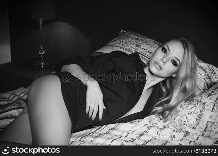 black and white ,beautiful blonde girl posing lying on bed with nude legs, unbuttoned blue shirt and fashion make-up. She is looking in camera with charming and relaxed expression ,