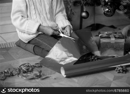 Black and white closeup image of cute girl cutting wrapping paper under Christmas tree at living room