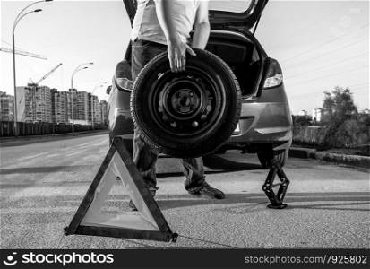 Black and white closeup photo of man carrying spare wheel against broken car