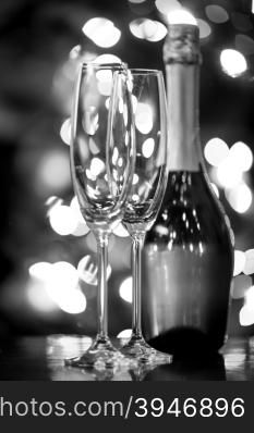Black and white closeup photo of two glasses and bottle of Champagne against Christmas lights