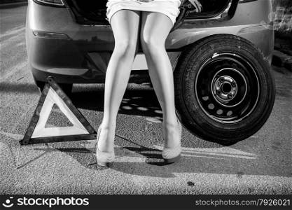 Black and white closeup photo of woman sitting on broken car near warning sign and spare wheel