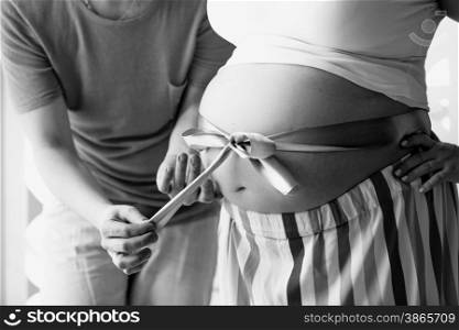 Black and white closeup photo of young man untying ribbon on wife&rsquo;s pregnant abdomen