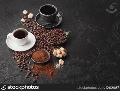Black and white cups of fresh raw organic coffee with beans and ground powder with cane sugar cubes with coffee tree leaf on black background.