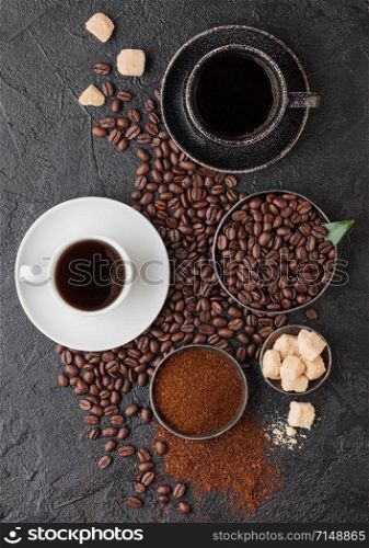 Black and white cups of fresh raw organic coffee with beans and ground powder with cane sugar cubes with coffee tree leaf on black background.