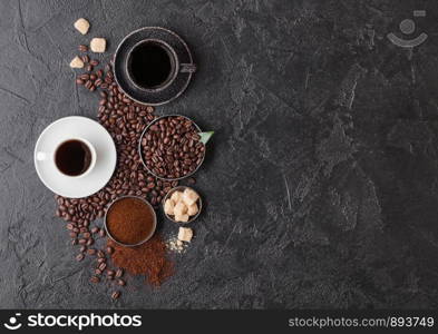 Black and white cups of fresh raw organic coffee with beans and ground powder with cane sugar cubes with coffee tree leaf on black background. Space for text