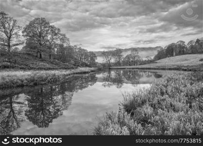 Black and white Epic Autumn landscape image of River Brathay in Lake District lookng towards Langdale Pikes with fog across river and vibrant woodlands