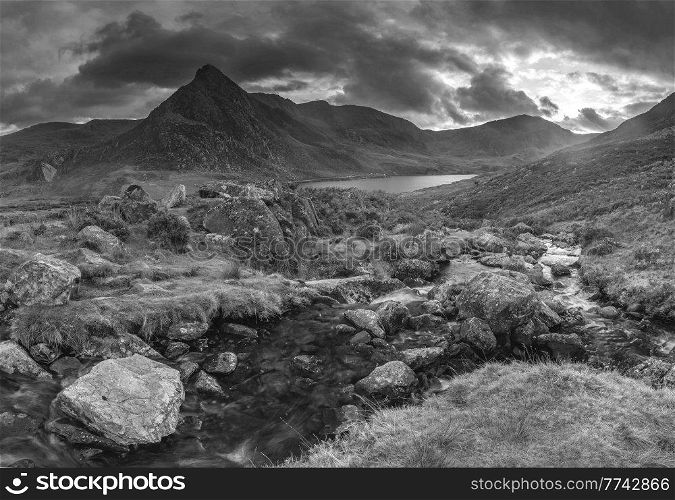 Black and white Epic dramatic Autumn sunset landscape image of Llyn Ogwen and Tryfan in Snowdonia National Park with stream and rocks in foreground