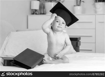 Black and white funny image of naked baby in diapers and graduation cap sitting on sofa